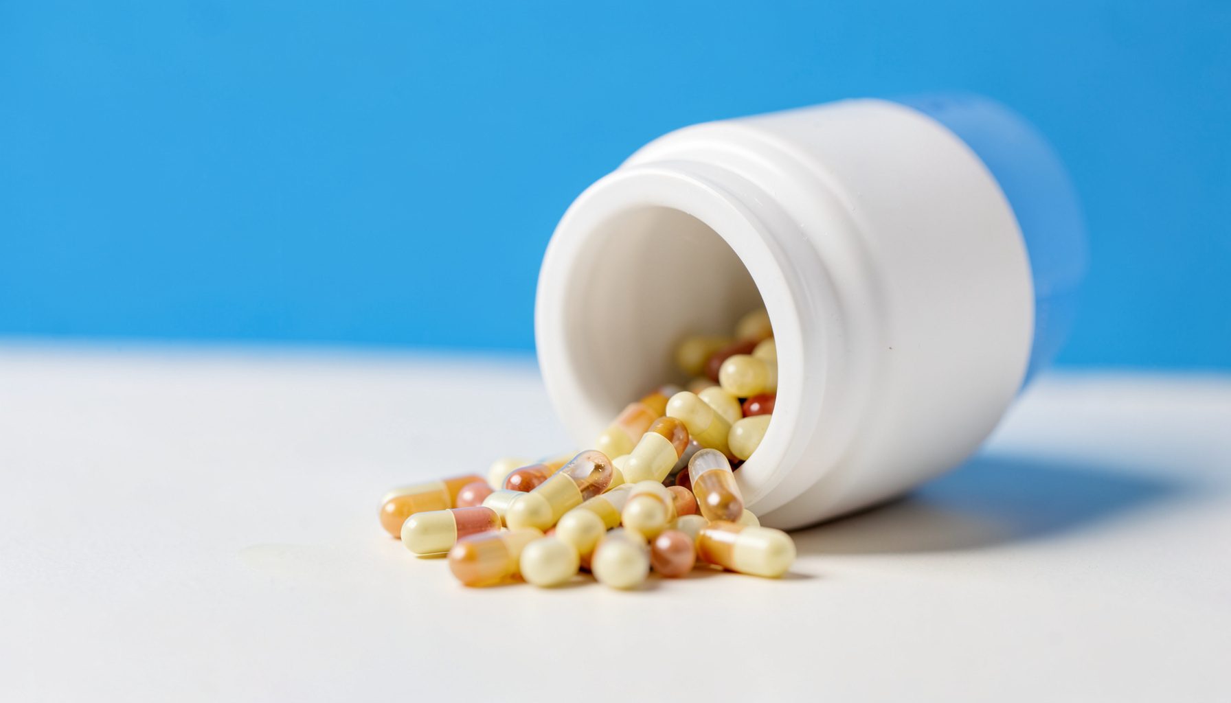 A close-up of weight loss capsules spilling from a white bottle onto a pale surface, soft diffused studio light.